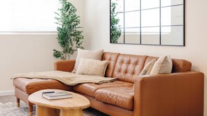 Ein braunes Ledersofa mit Decke und Kissen, ein runder Couchtisch mit einem Buch und Fernbedienung sowie ein großes gerahmtes Bild an der Wand.