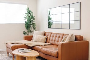 Ein braunes Ledersofa mit Decke und Kissen, ein runder Couchtisch mit einem Buch und Fernbedienung sowie ein großes gerahmtes Bild an der Wand.
