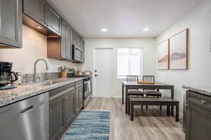 A modern kitchen with granite countertops, stainless steel appliances, and a dining area with a wooden table and chairs. - Coastal 2BR | Walk to Ocean Beach & Cafés (San Diego)