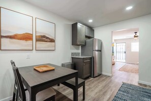 A modern kitchen with a dining table, two framed artworks, a refrigerator, and a view into another room. - Coastal 2BR | Walk to Ocean Beach & Cafés (San Diego)