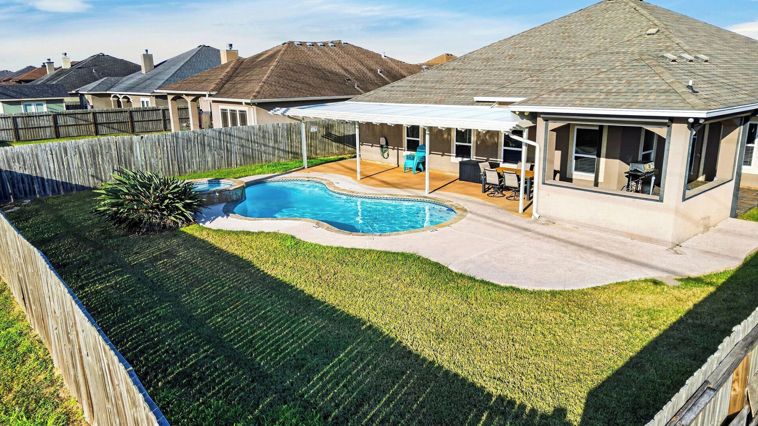 A backyard with a swimming pool, a covered patio area, and a wooden fence.