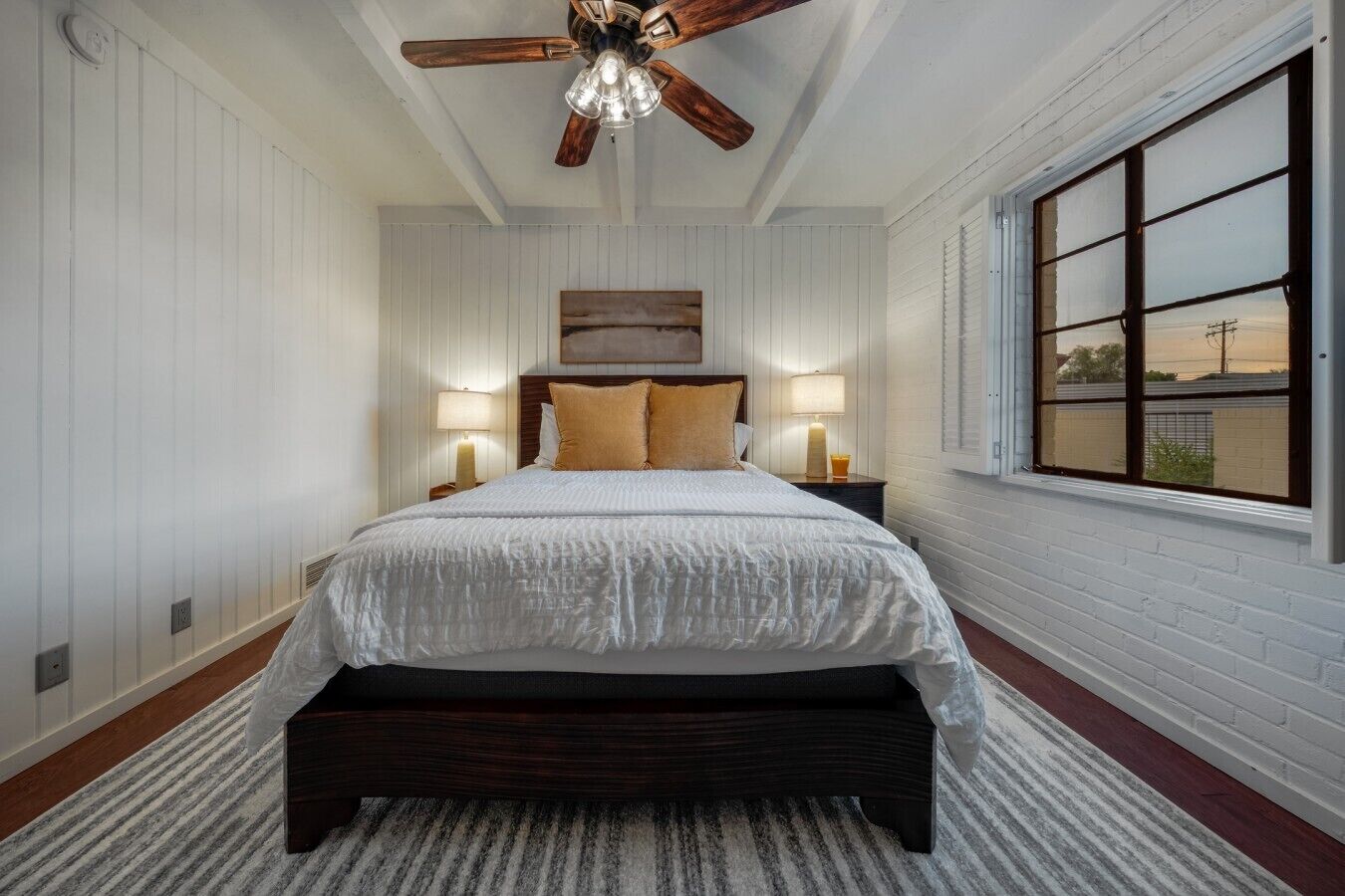 A bedroom with a large bed, two bedside tables with lamps, a ceiling fan, and a window with blinds.