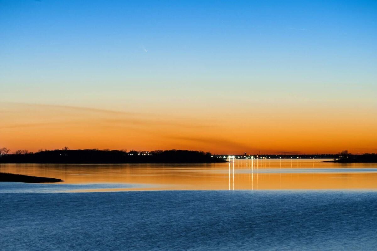 Água calma refletindo um céu ao pôr do sol, com uma linha de costa distante.