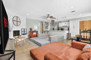 A modern living room with a sofa, ottoman, and a coffee table. There is a fireplace, a ceiling fan, and a kitchen area in the background. - Poolside Escape | Game Area | King Beds | Arcades (The Colony)