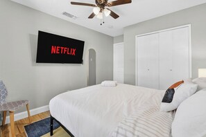 A bedroom with a bed, a TV mounted on the wall, a ceiling fan, and a door. - Poolside Escape | Game Area | King Beds | Arcades (The Colony)