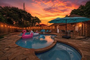A backyard with a swimming pool, inflatable flamingo float, and a brick building with outdoor seating. - Home Theater - Game Room - Pool - Hot Tub - Golf (Plano)