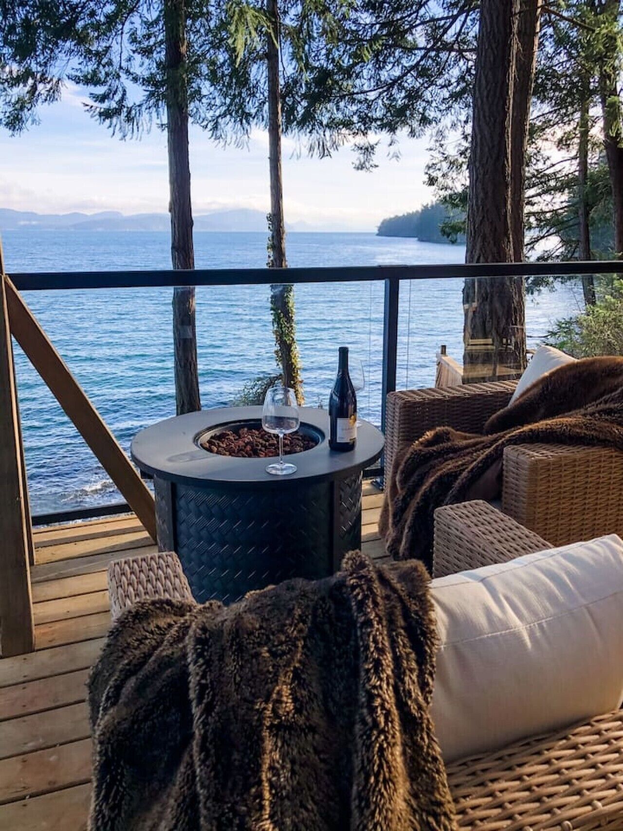 A deck with a round table, a bottle, and a glass, overlooking a body of water and trees.