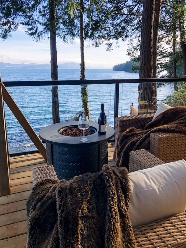 Una terraza con una mesa redonda, una botella y un vaso, con vista a un cuerpo de agua y árboles.