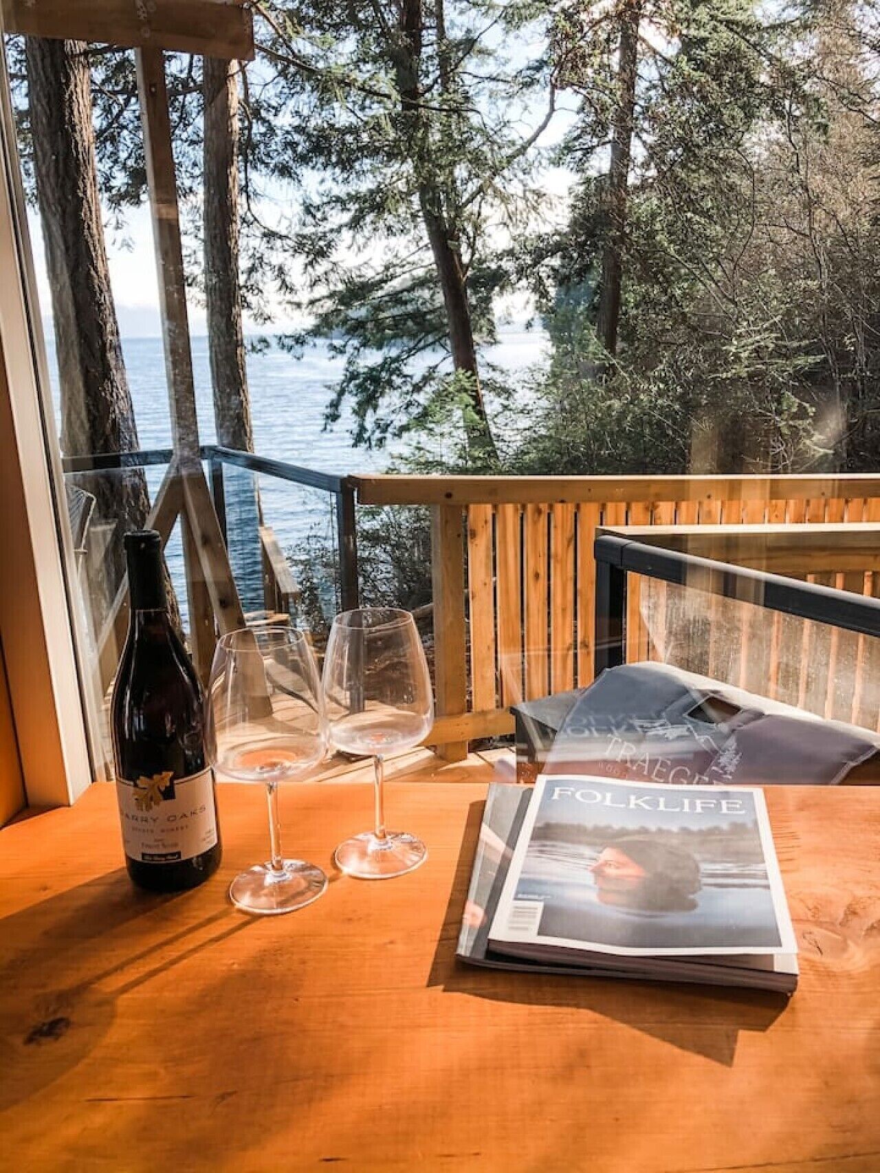 Une table en bois, une bouteille de vin, deux verres à vin et un magazine sont posés sur un balcon donnant sur une forêt et un plan d’eau.