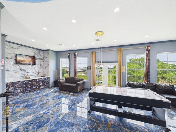 A spacious room with a marble floor, a pool table, a sofa, and large windows offering a view of greenery.