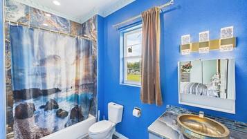 A bathroom with a blue wall, a shower curtain with a seascape design, a white toilet, a window, and a vanity with a mirror and sink.