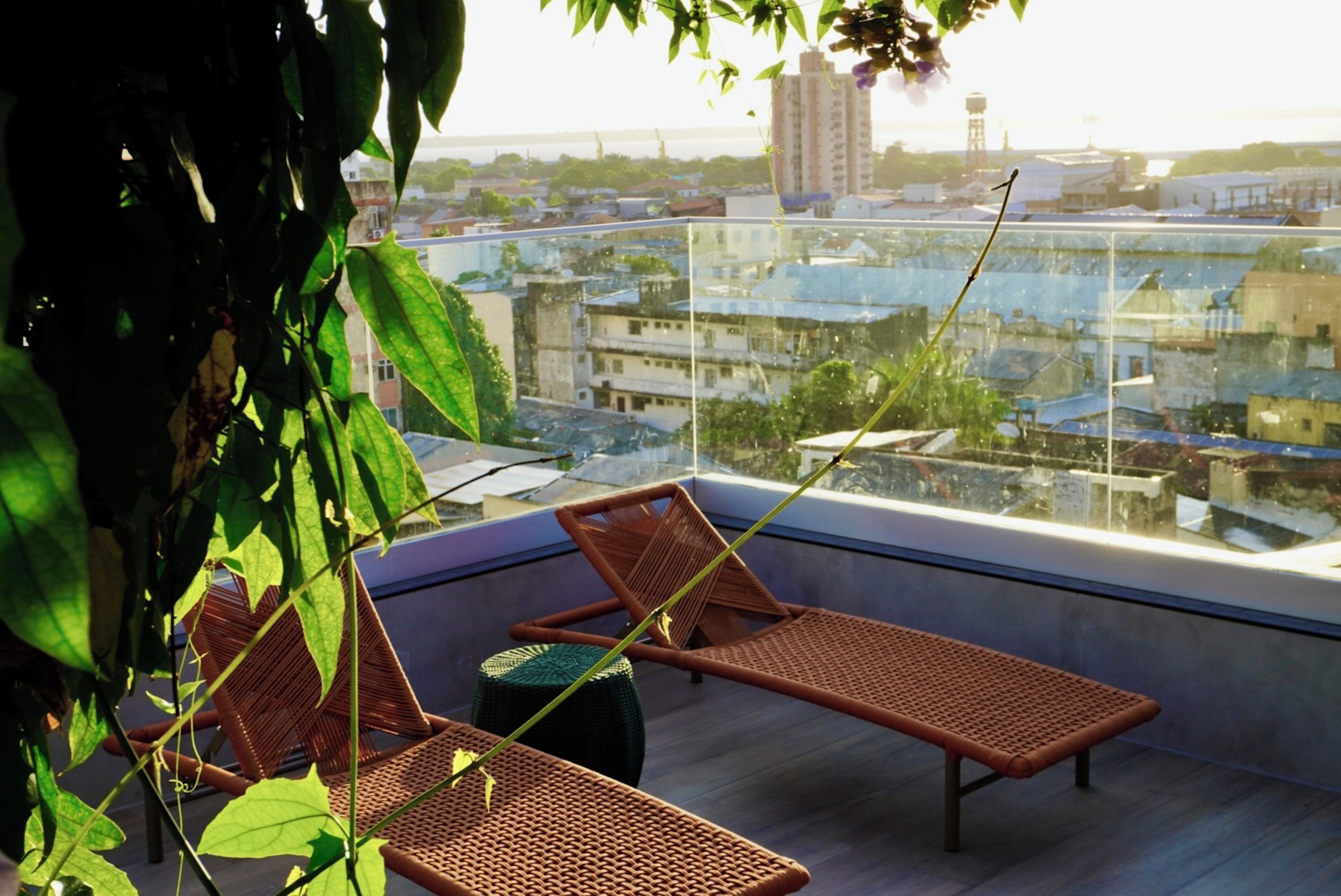 Varanda com duas poltronas de vime e uma pequena mesa, com vista para a paisagem urbana com edifícios e um tanque de água.