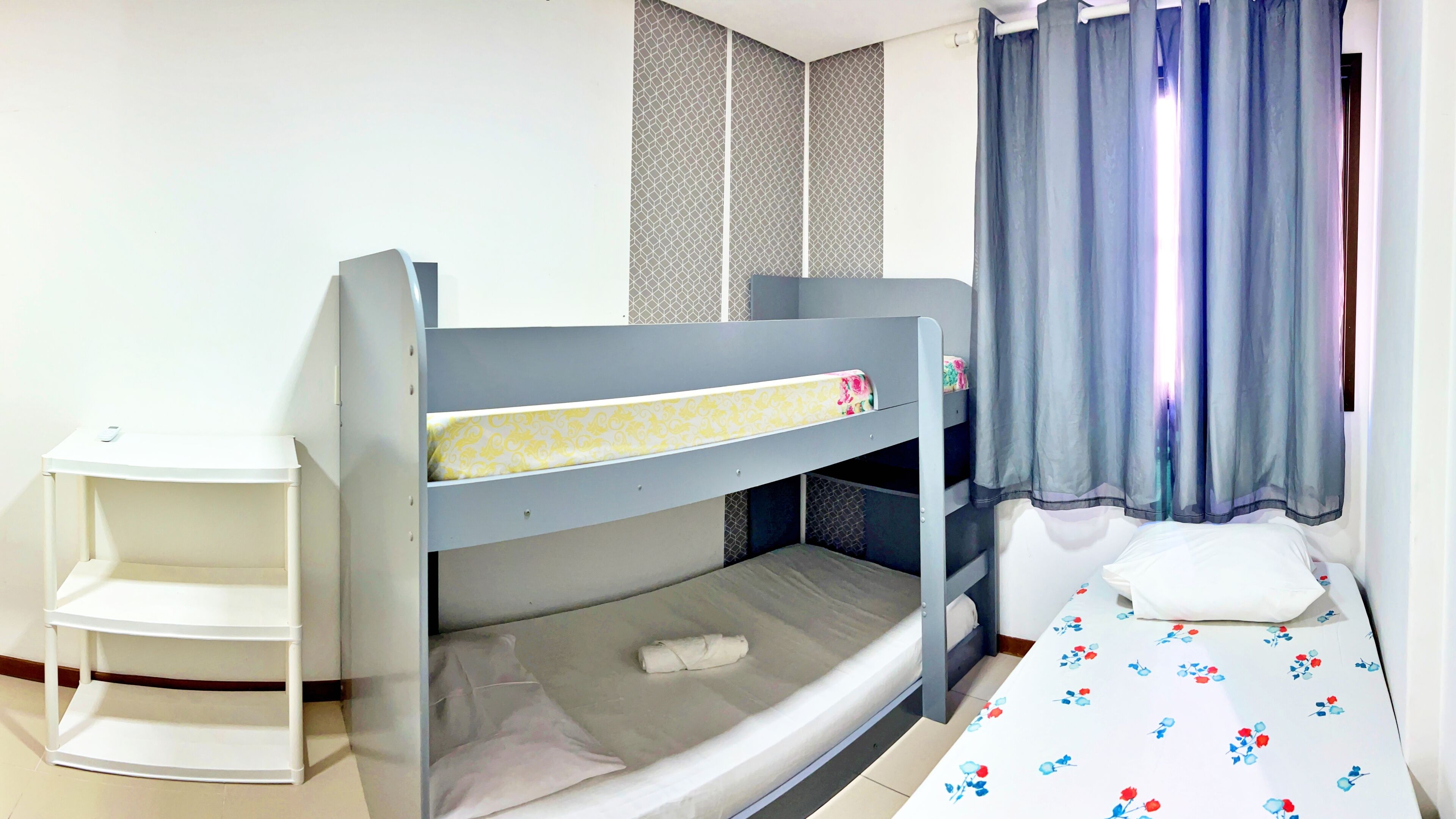 Cama litera con un colchón blanco y una litera inferior con un colchón gris. Hay una mesita de noche blanca con un objeto pequeño encima.