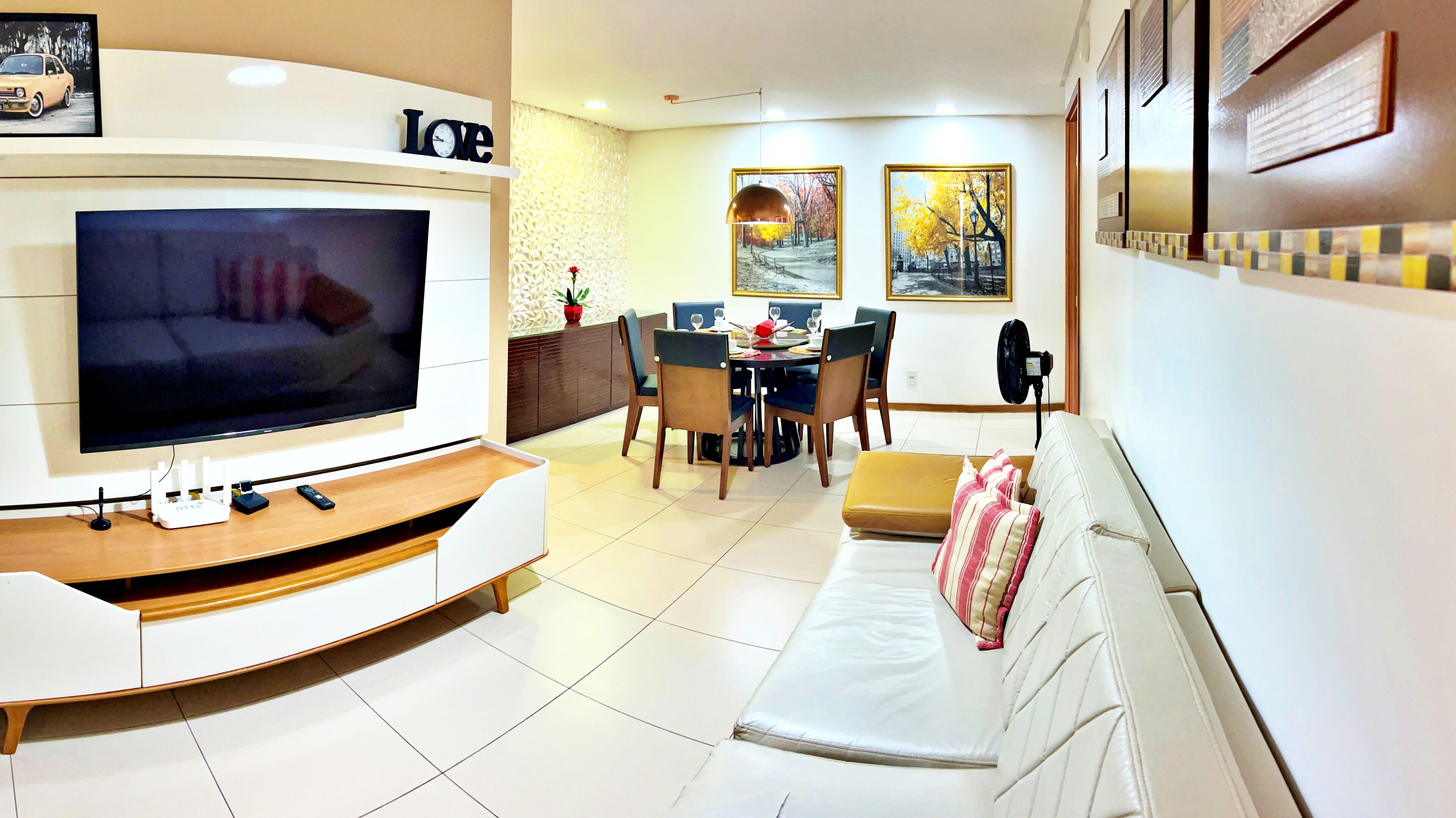 Una sala de estar moderna con televisor de pantalla plana, una zona de comedor con mesa y sillas, y un sofá con cojín.