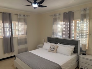 Ein Schlafzimmer mit einem Bett, Nachttischen, einem Deckenventilator und Fenstern mit Vorhängen.