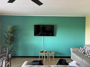 Ein Zimmer mit türkisfarbenen Wänden, einem an der Wand montierten Flachbildfernseher, einem Esstisch mit Stühlen, einem Sofa und einem kleinen Beistelltisch.