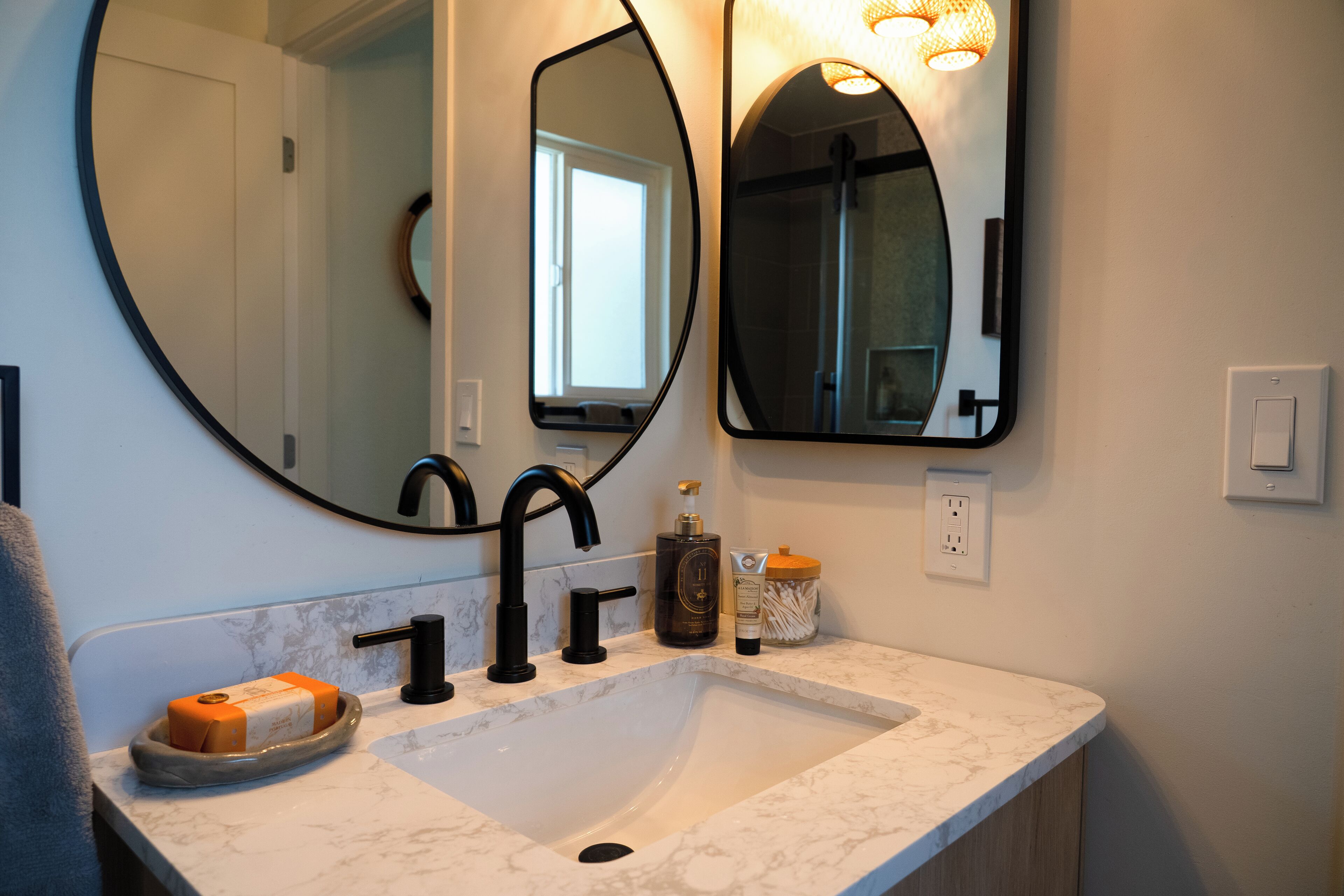 A bathroom with a white marble sink, black faucets, a round mirror, and a bottle of lotion.