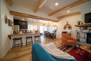 Ein modernes Wohnzimmer mit blauem Sofa, Holzbalken, einem Steinofen und einer Küchenzeile mit Bar.