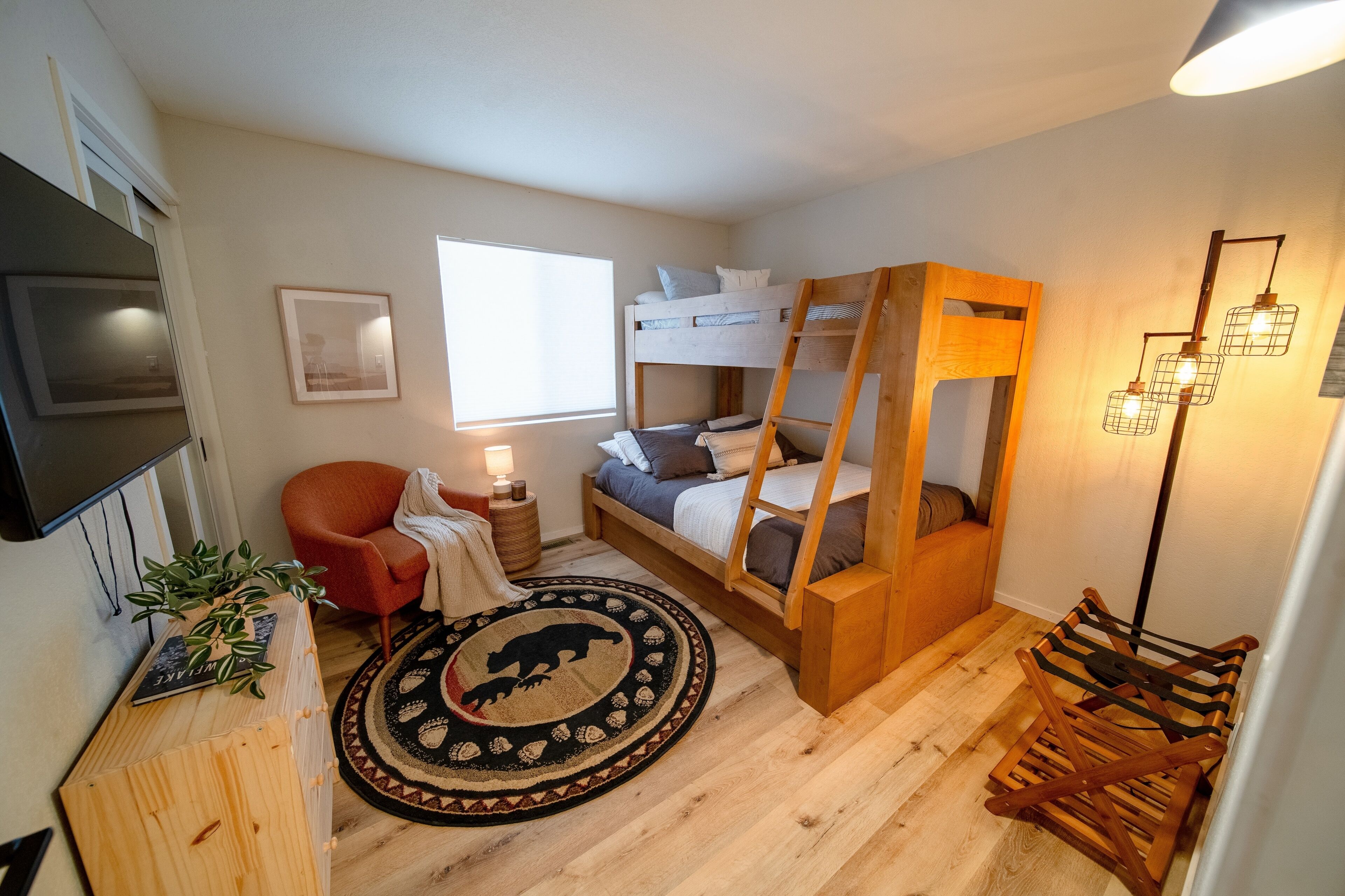 Et lille værelse med køjeseng, enkeltseng, rød stol, lille bord med lampe, plante, rund måtte og en træstige.