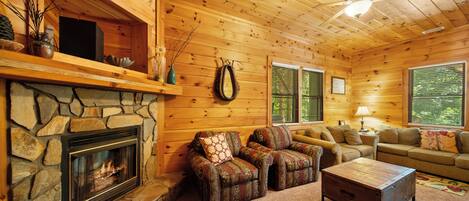 A cozy cabin interior with a stone fireplace, wooden walls, a stone hearth, a sofa set, and a ceiling fan.