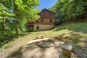 Eine Holzhütte mit einer Terrasse, umgeben von Bäumen und einem Kiesplatz mit Feuerstelle.