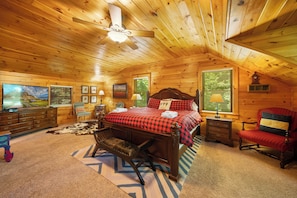 Une chambre confortable avec un plafond en bois, un lit recouvert d’une couette à carreaux rouges, une télévision et une grande fenêtre avec des stores.