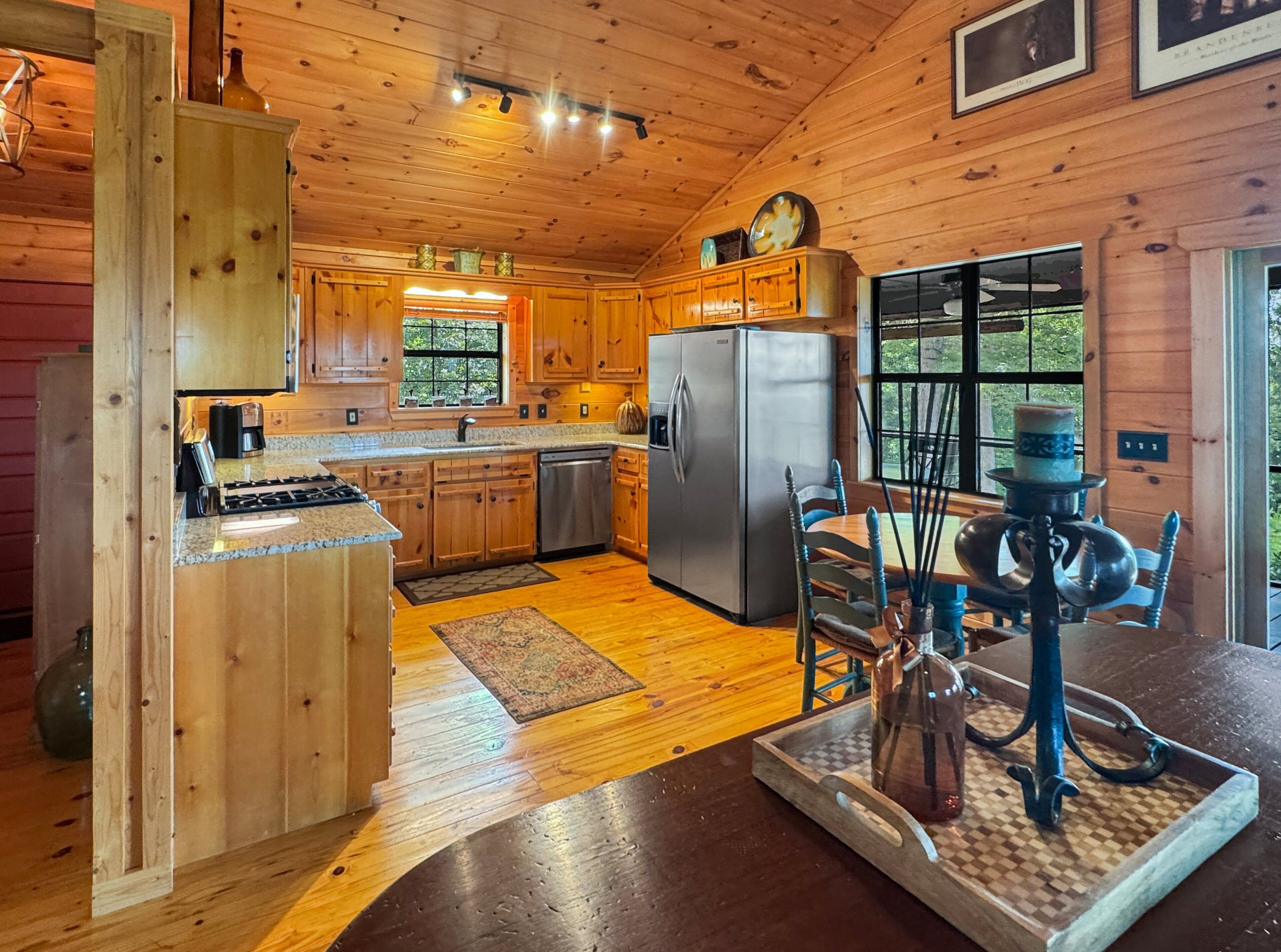 A wooden cabin interior with a kitchen, dining area, and a large window.