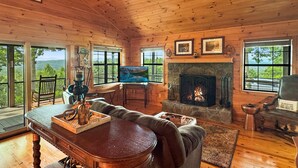 A cozy cabin interior with a stone fireplace, wooden furniture, and large windows offering a view of the outdoors. - Southwind Ridge - Mountain View | Screened porch | Fireplace (Morganton)