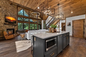A spacious kitchen with a large island, a stone fireplace, and a view of the outdoors through large windows. - Hope Ridge Manor - Pet Friendly | Firepit | Hot Tub | Game Room (Blue Ridge)