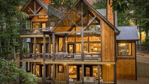 A multi-story wooden cabin with a balcony, large windows, and a visible interior with seating. - Hope Ridge Manor - Pet Friendly | Firepit | Hot Tub | Game Room (Blue Ridge)