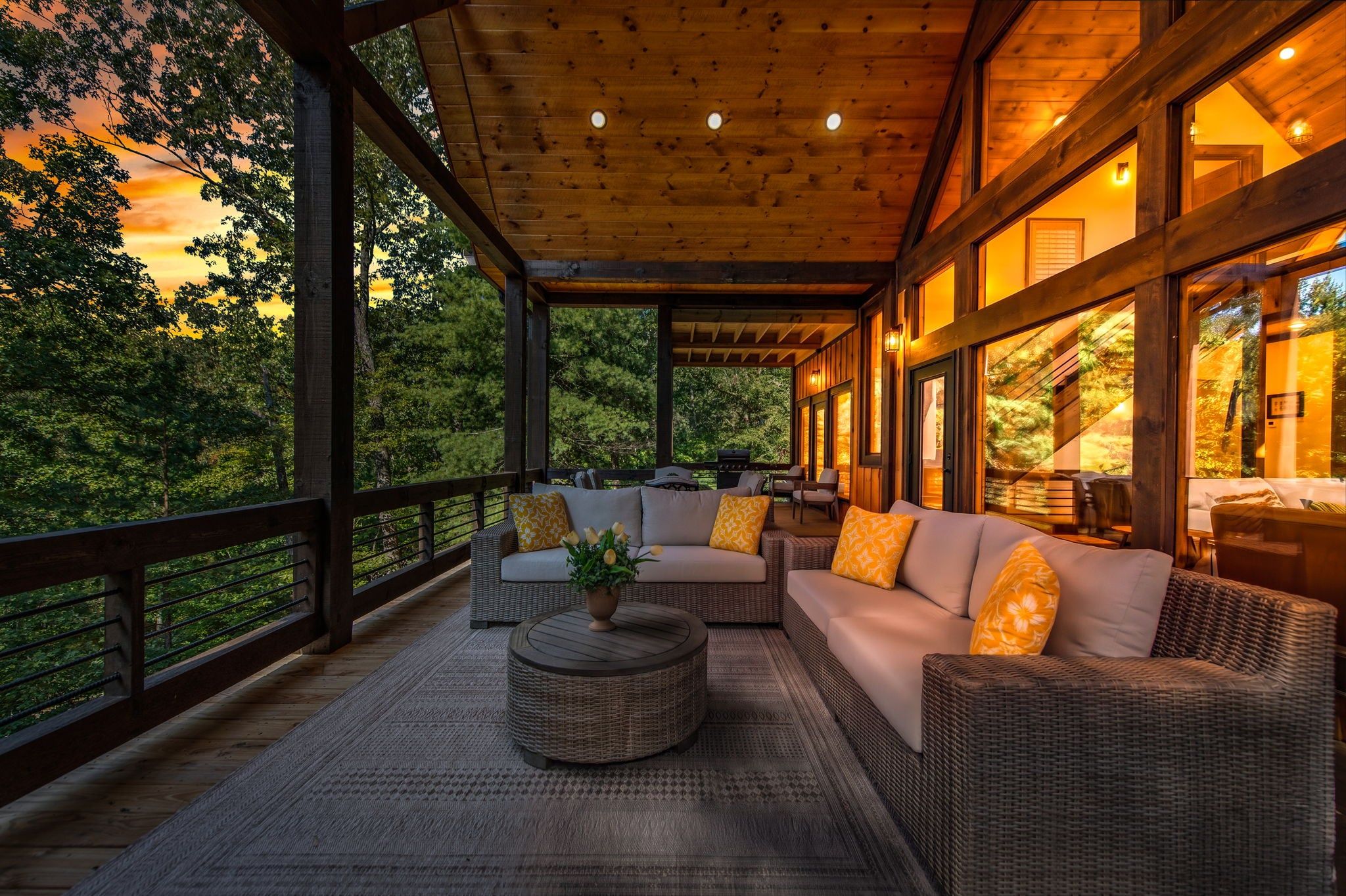 A covered outdoor seating area with wicker furniture and a round coffee table, overlooking a forested landscape.