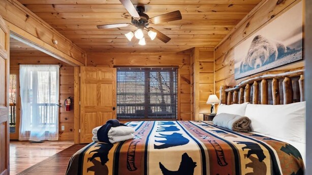 Una camera in una casetta di legno con un letto, una ventola a soffitto, una finestra con tende e un quadro di un orso al muro.
