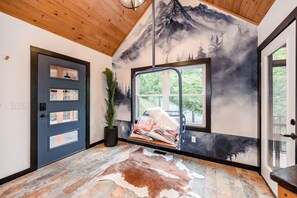 Ein Zimmer mit Wandmalerei von Bergen, einer Holzbalkendecke, einer Tür mit mehreren Feldern, einem Fenster mit Blick auf Grünflächen und einem Kuhfellteppich.