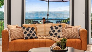 Un sofá de cuero con cojines estampados, una mesa de centro con libros y vistas a un paisaje montañoso a través de ventanas amplias.