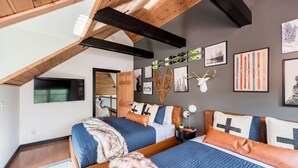 Un dormitorio con techo inclinado, vigas de madera, dos camas con ropa de cama azul y arte en la pared, incluyendo esquíes y una cabeza de ciervo.