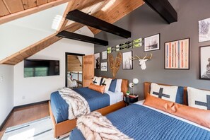 A bedroom with a sloped ceiling, wooden beams, two beds with blue bedding, and wall-mounted artwork including skis and a deer head. - R&R Chalet | Ski View | Hot Tub | Theater | Sauna (McGaheysville)