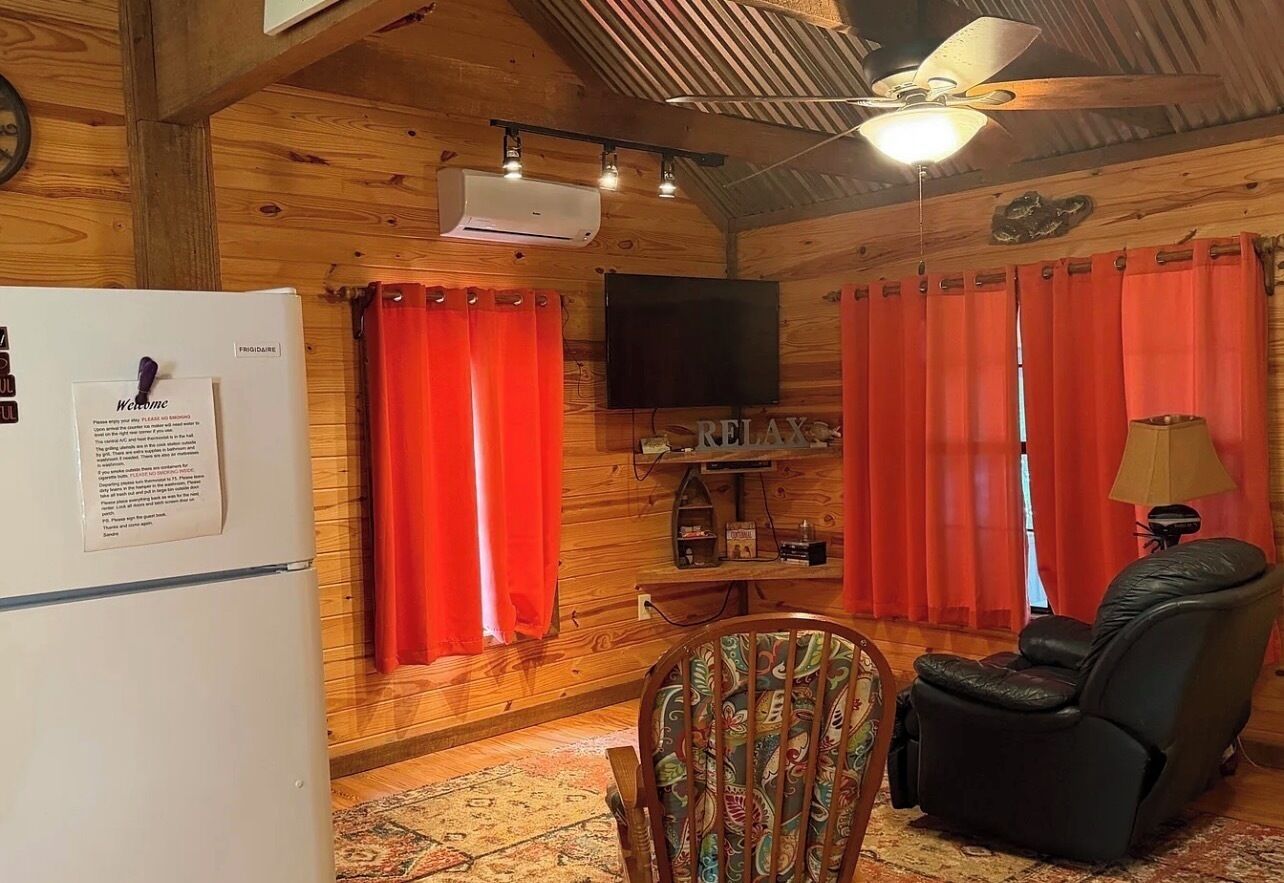 A cozy cabin interior with a wooden ceiling, a ceiling fan, a mounted TV, and a refrigerator.