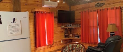 A cozy cabin interior with a wooden ceiling, a ceiling fan, a mounted TV, and a refrigerator.