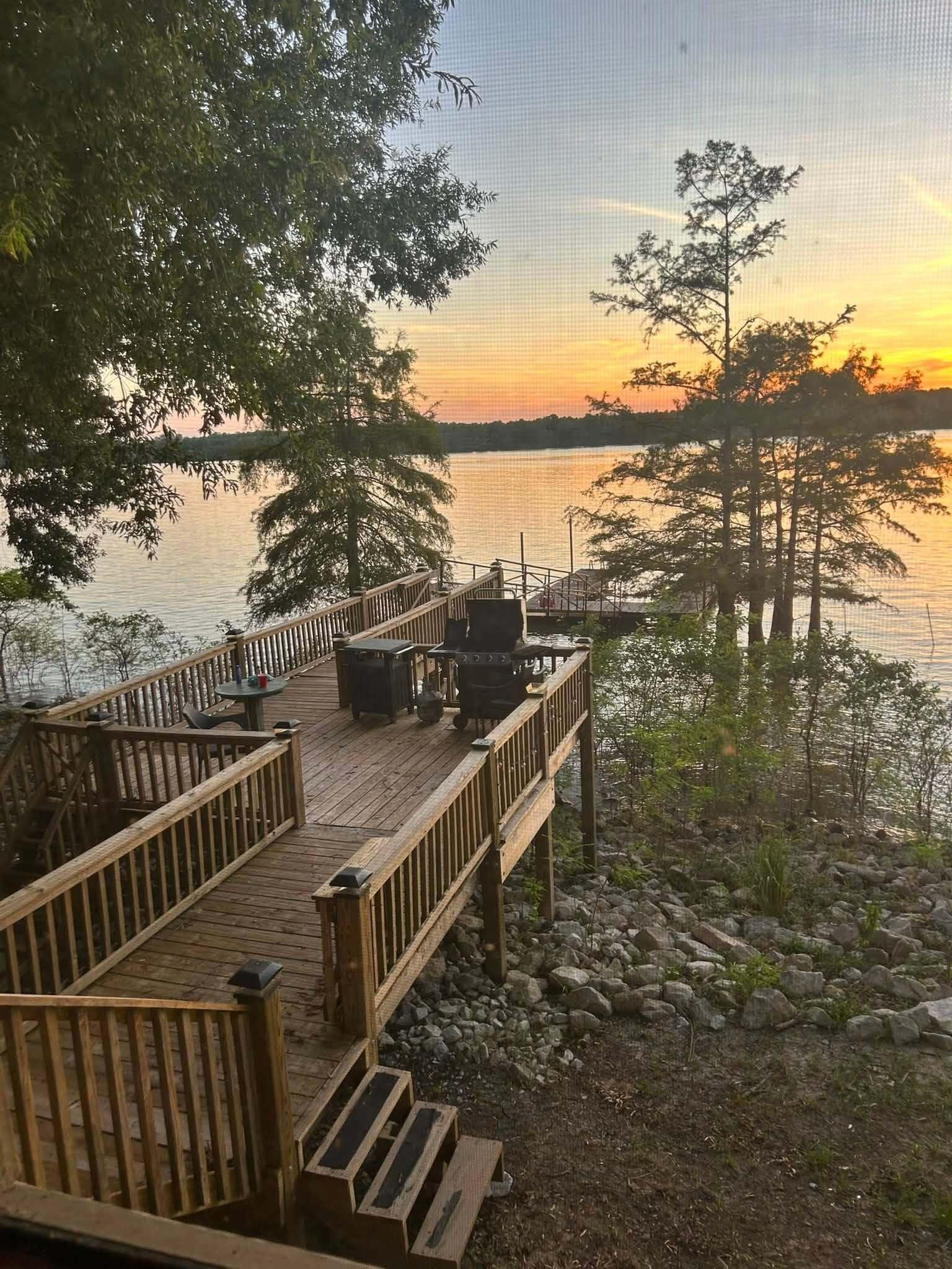 A wooden deck with outdoor seating and a grill, overlooking a lake with trees and a sunset.