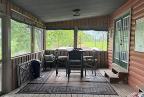 A covered porch with a table and chairs, a view of the outdoors, and a wooden floor.