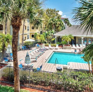 A pool area with lounge chairs and umbrellas, surrounded by palm trees and a building with a tiled roof. - Bright Beach Condo - Minutes to Sand and Sunsets (Clearwater)