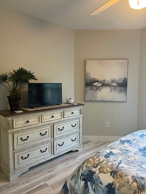 Ein Schlafzimmer mit Bett, Kommode mit Schubladen, Fernseher auf der Kommode, Topfpflanze und einem Bild an der Wand.