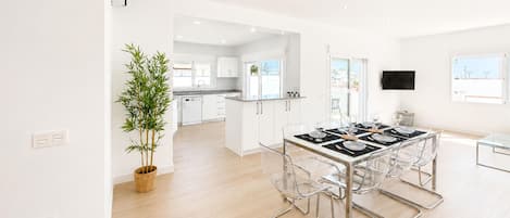 A modern kitchen with white cabinets, a dining area with a glass table and chairs, and a large window providing natural light.