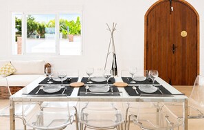 Un coin repas avec une table dressée pour six personnes, des chaises transparentes, une porte en bois et une fenêtre donnant sur un paysage verdoyant.