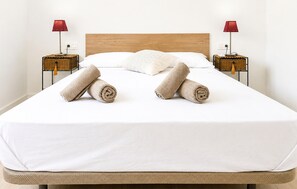 Un lit bien fait, avec une tête de lit en bois, du linge de lit blanc et des serviettes enroulées.