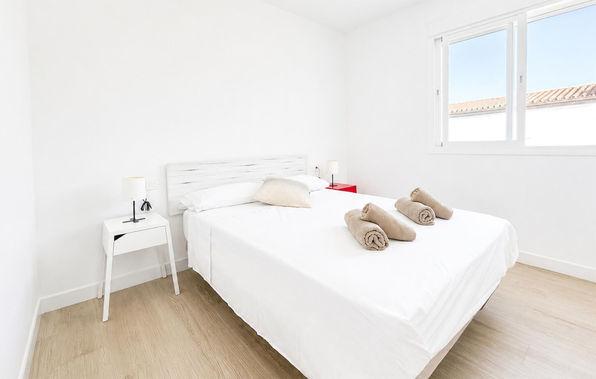 Une chambre à coucher minimaliste avec un lit simple, une table de chevet, une petite table d’appoint rouge et une fenêtre donnant sur un immeuble.