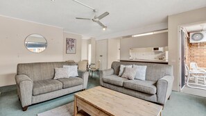 Un salon avec deux canapés gris, une table basse en bois, un ventilateur de plafond et, au fond, une cuisine.