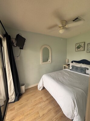 Ein Schlafzimmer mit einem Bett, einem Deckenventilator, einem an der Wand befestigten Fernseher, einem Spiegel und einem Fenster mit Vorhängen.