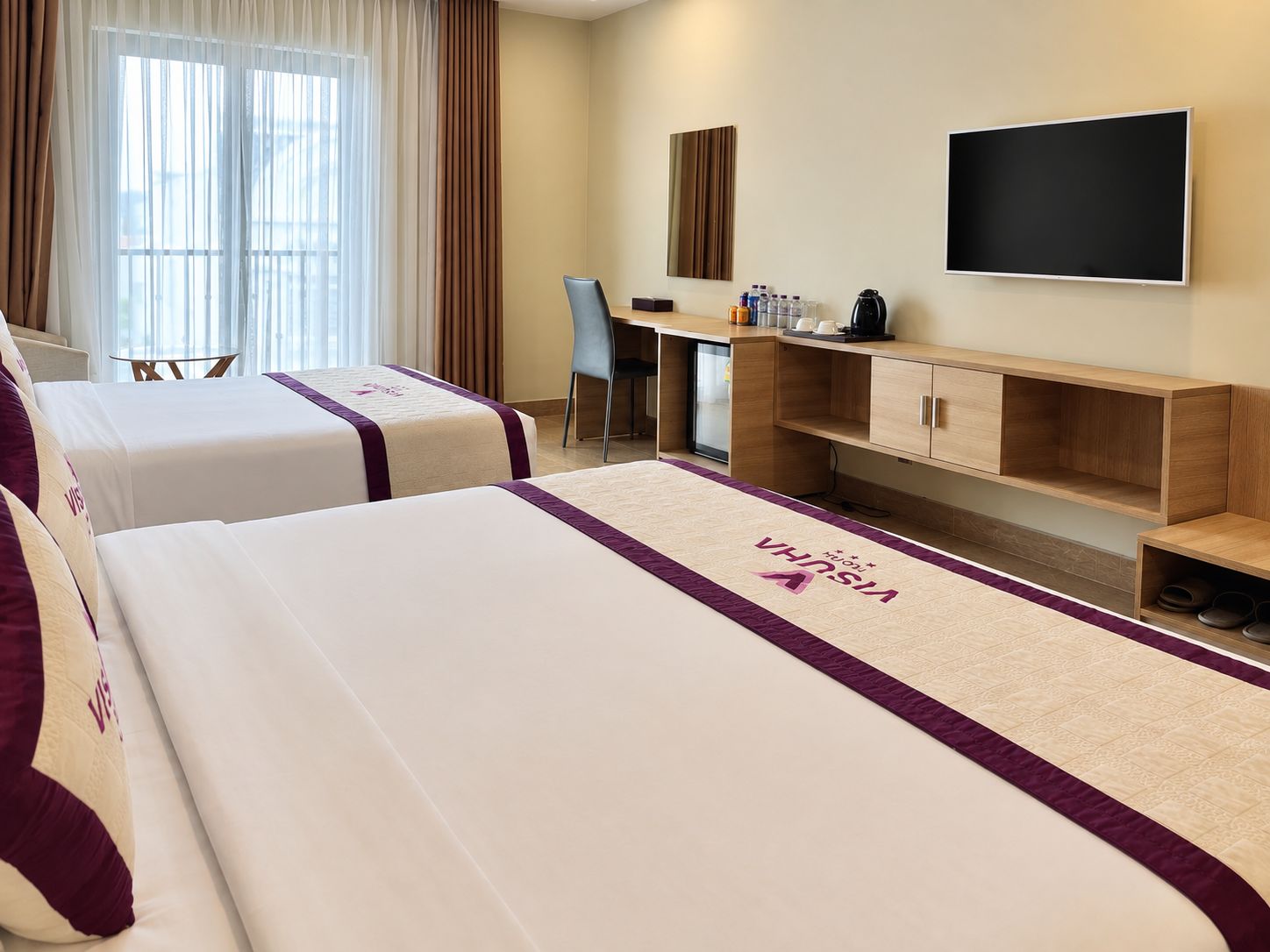 A hotel room with two beds, a desk, a chair, a TV, and a minibar.