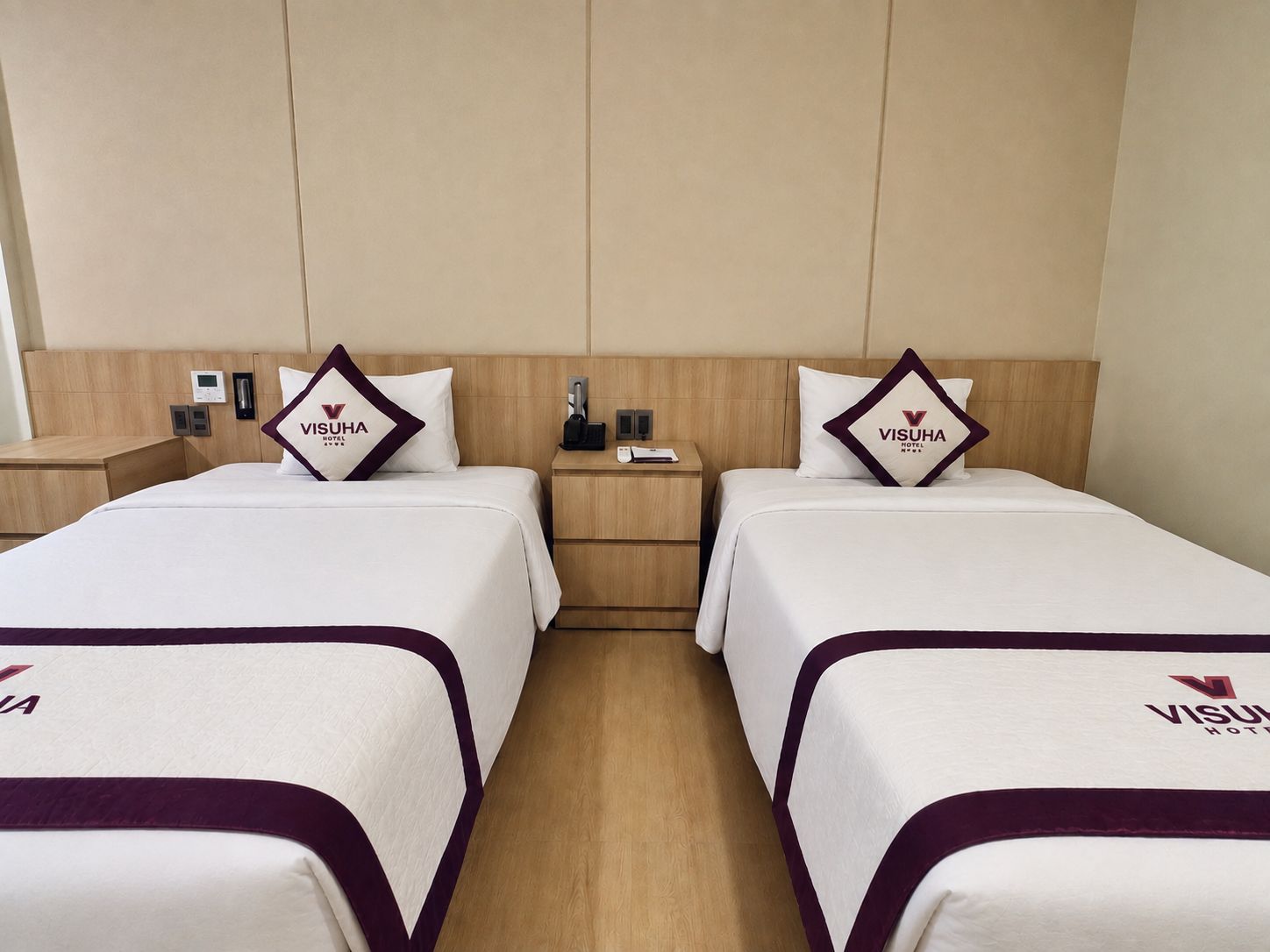 A hotel room with two beds, each with a pillow featuring the hotel logo. There are bedside tables with electronic devices and a wall-mounted phone.